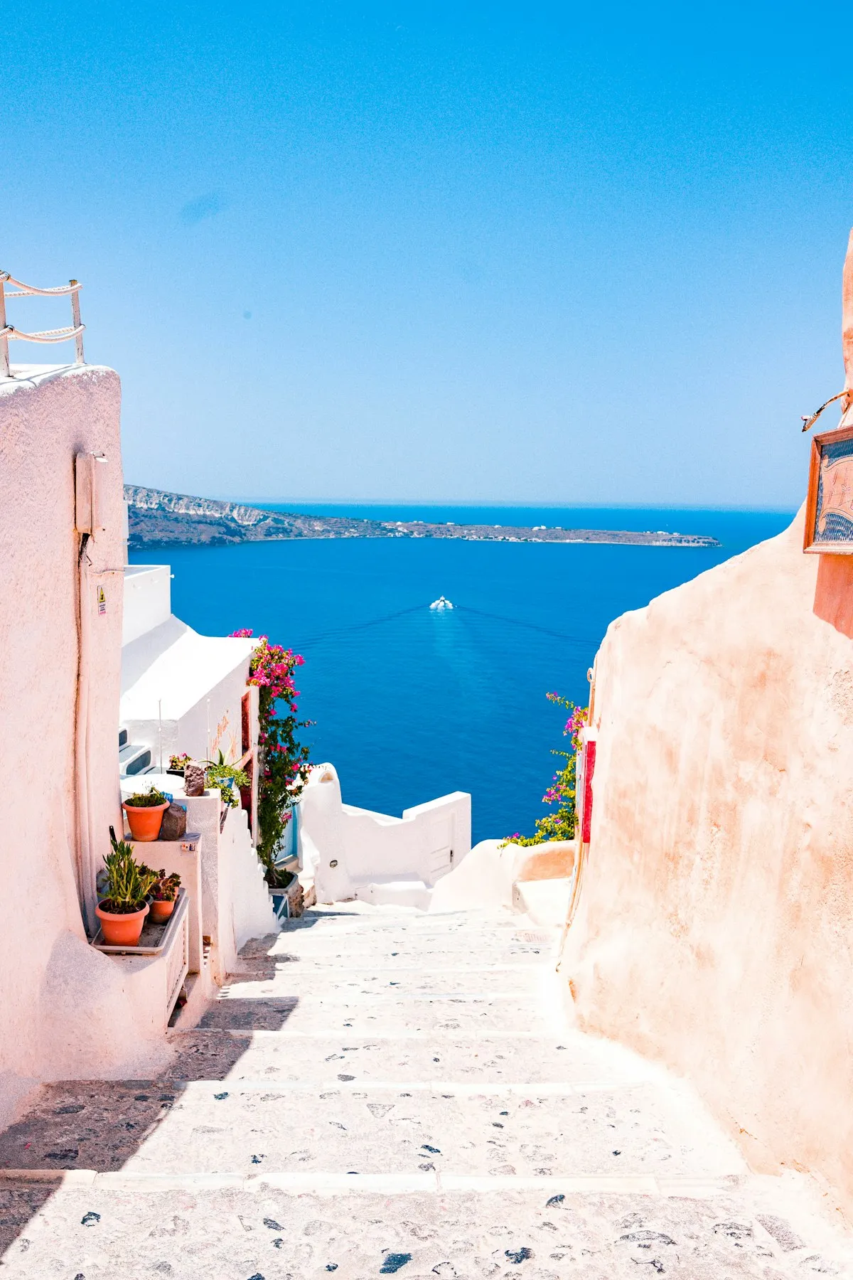 Whitewashed steps with potted flowers leading down between pink walls to a deep blue sea